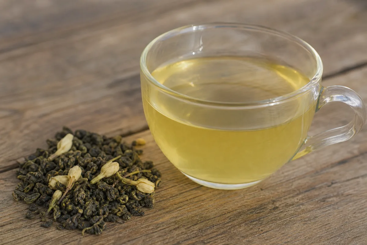 A clear glass cup of pale golden-green jasmine tea beside a small pile of rolled dark green tea leaves with dried jasmine flower buds