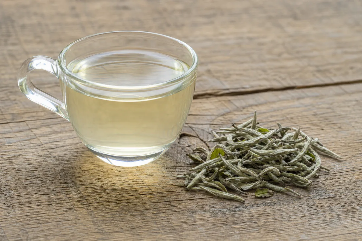 A clear glass cup of very pale straw-yellow white tea beside a small pile of dried silvery-green white tea buds and pale young leaves